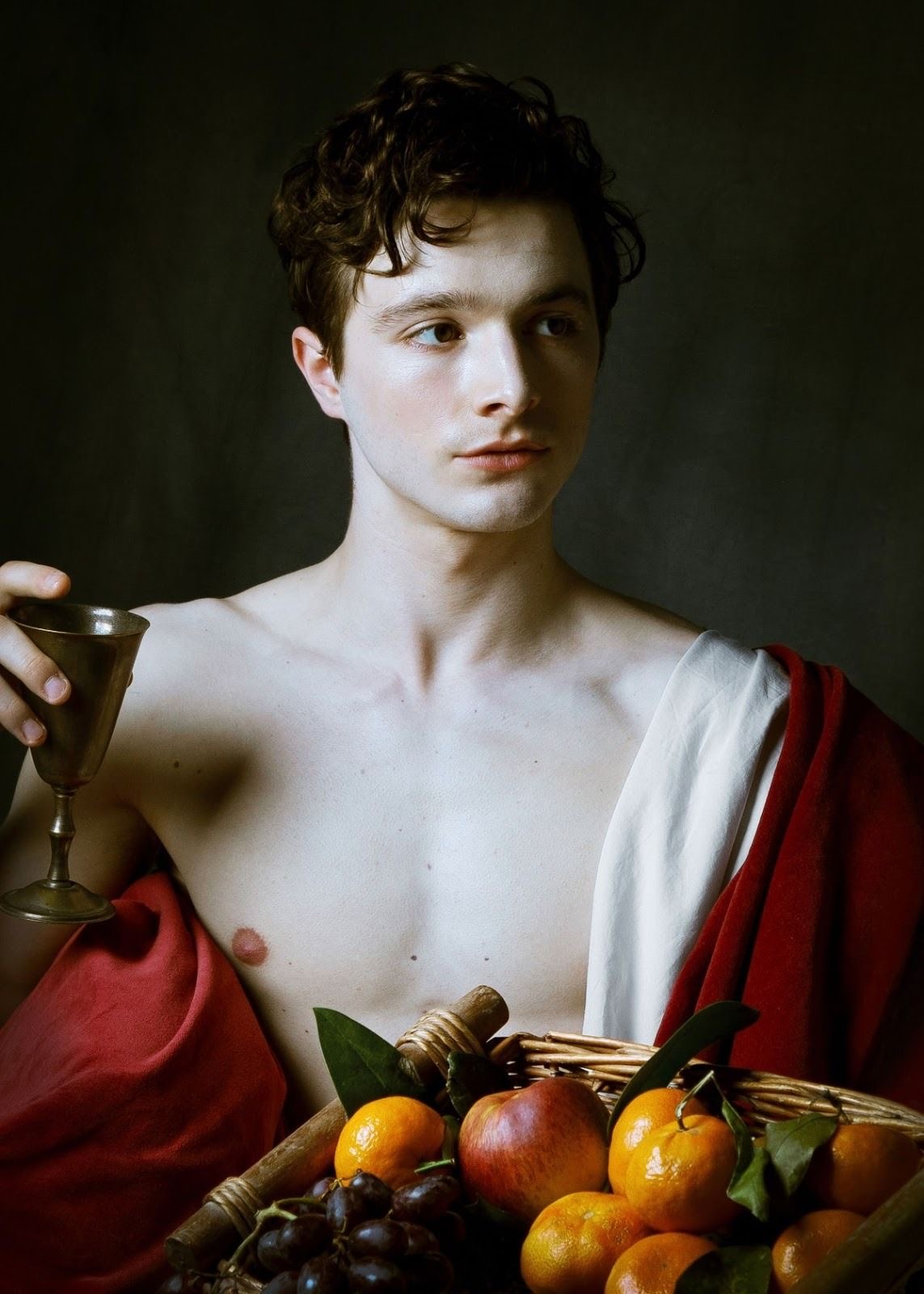 a young man, shirtless and draped in red and white fabric, poses for a portrait in the style of a classical painting. he holds a metal goblet in his right hand and gazes off to the side.  in the foreground, a woven basket overflows with a vibrant arrangement of grapes, apples, and oranges. the lighting is dramatic, highlighting the subject's musculature and the textures of the fruit and fabric. the dark background emphasizes the subject and adds to the painting's overall sense of drama and timelessness.