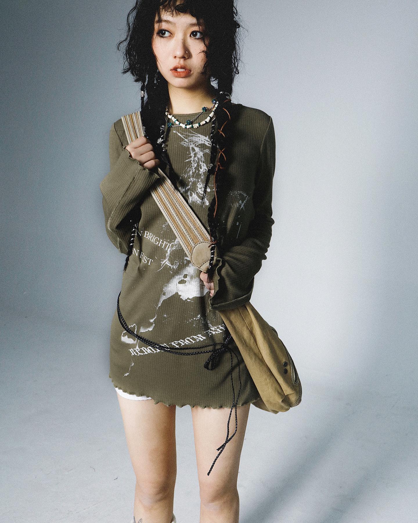 a young woman with dark, short hair poses in a studio against a grey background. she is wearing a green longsleeved ribbed shirt with a graphic print and text, a pair of short white shorts underneath, a necklace, and a crossbody/tote bag. she is looking away from the camera.
