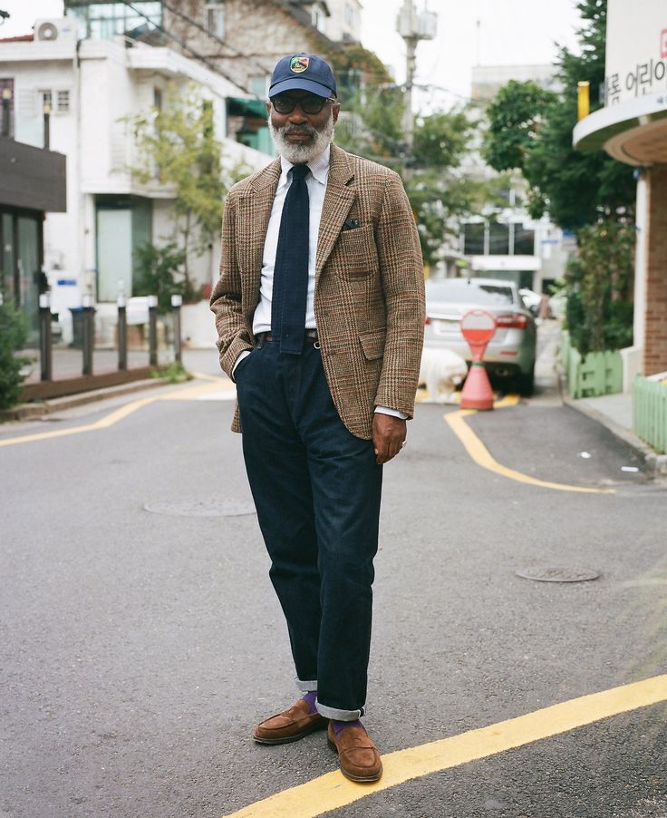 n older man with a grey beard and white hair is standing on a city street. he's wearing a brown plaid blazer, dark blue jeans, brown suede loafers with purple socks, and a baseball cap. his style is casual yet elegant. the photo appears to have a vintage feel. the background shows a street in what looks like a korean city.