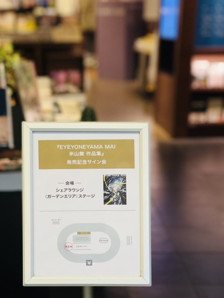 the image shows a white sign within a bookstore setting. the sign, slightly angled towards the viewer, announces a book signing event for the art book "eyeyoneyama mai" by artist 米山舞 (yoneyama mai). the sign features japanese text, a black and white illustration from the book, and a small floor plan indicating the signing location within the store. the bookstore background is blurred, suggesting a bustling atmosphere.