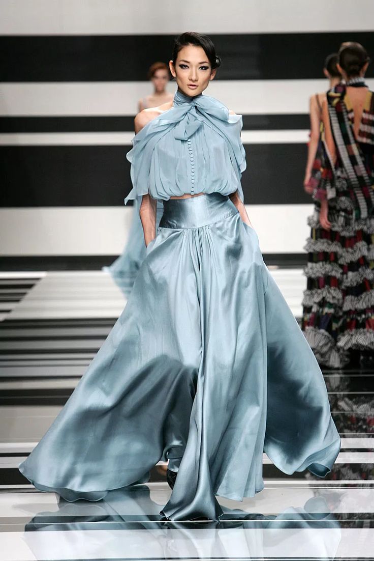 a model walks the runway in a light blue satin gown featuring a crop top with a highnecked bow and voluminous, highwaisted wideleg pants. the background is a black and white striped pattern. other models are visible in the background.
