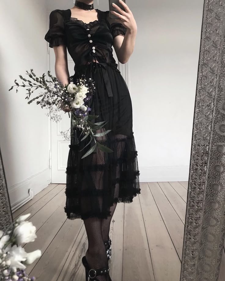 a person is taking a selfie in a mirror, dressed in a black outfit consisting of a blouse with a sweetheart neckline and puff sleeves, and a long, tiered skirt. they are also wearing a black choker, fishnet stockings, and highheeled shoes with straps. they are holding a bouquet of white and purple flowers. the mirror has an ornate silver frame. the background shows a white wall and wooden floor.