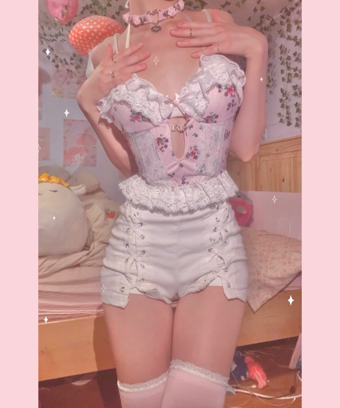 the image shows a young woman wearing a delicate pink and white lingerie outfit. the outfit consists of a floral corset top with lace detailing, matching ruffled trim, and white laceup shorts.  she is also wearing a delicate necklace and rings. the overall style is very kawaii and feminine, with a pastel pink and white color scheme. the setting appears to be a bedroom with soft lighting and pink decor. the woman is posed in a way that accentuates the clothing, and the overall aesthetic is soft, romantic and playful.  she's also wearing light pink thighhigh socks.
