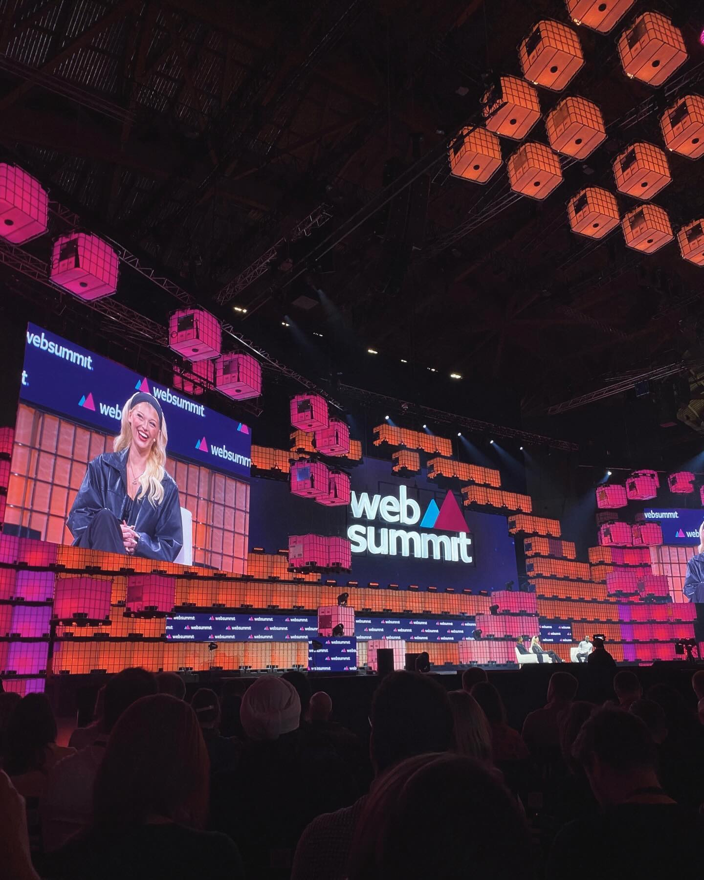 a wide shot of a web summit event, featuring a stage with a large screen showing a woman speaking, the "web summit" logo, an audience, and hanging illuminated cubeshaped decorations.
