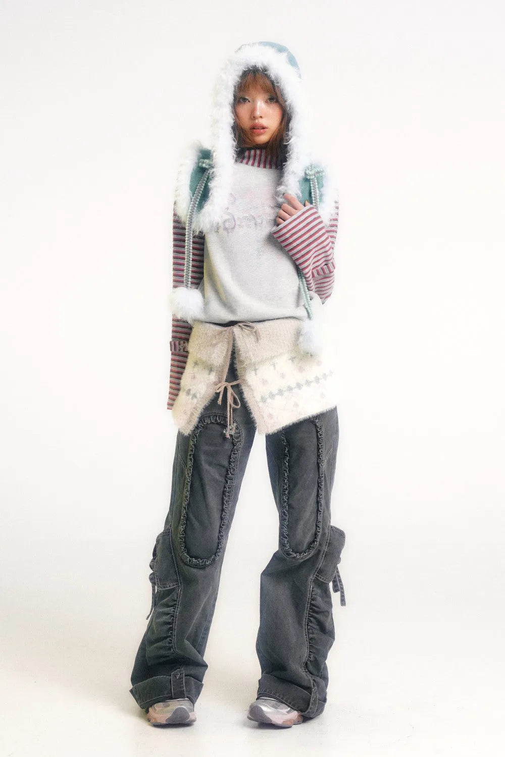 fullbody studio shot of a young asian woman modeling a winter fashion ensemble. she's wearing a hooded hat with white fur trim, a teal vest over a striped longsleeved shirt and a lightcolored sweater.  she also sports a knit belt and dark gray, patched jeans with cinched ankles, and is wearing sneakers.  the background is plain white.