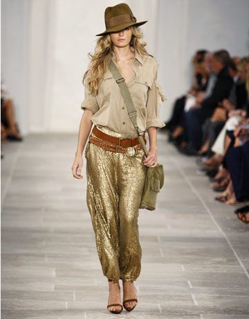 the image shows a fashion model walking on a runway. she's wearing a stylish outfit that blends safari and bohemian elements.  the outfit includes a khakicolored buttondown shirt, gold sequined haremstyle pants, a brown leather belt, a brown fedora hat, and brown leather sandals. she carries a green canvas shoulder bag. the model's blonde hair is styled in loose waves. the background shows other models and audience members blurred to emphasize the main model. the overall style is sophisticated and glamorous, indicative of a highfashion runway show, likely from a brand like ralph lauren, given the aesthetic.