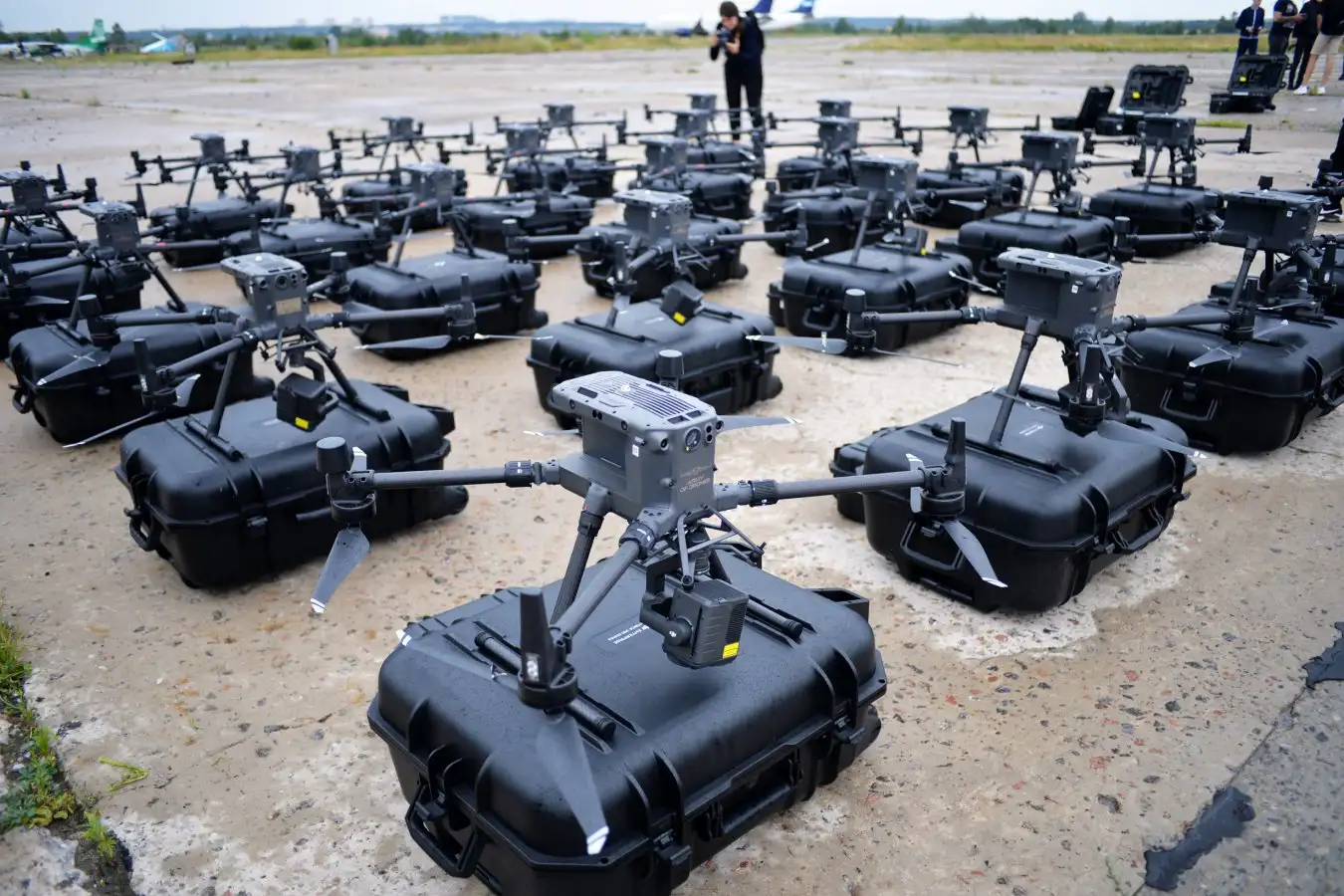 a large number of drones are lined up on the tarmac. they appear to be in storage containers and are all black. a few people can be seen in the background as they are likely operating the drones.