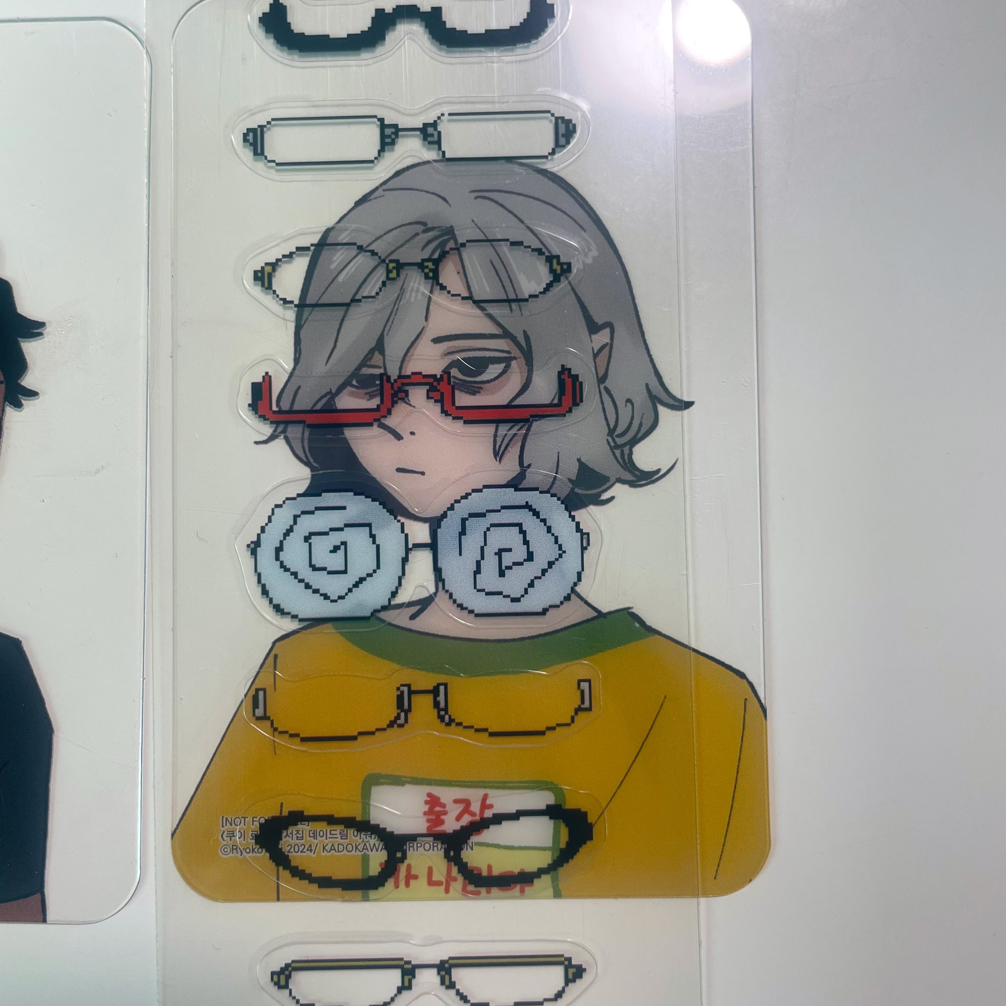 loseup view of a transparent sticker sheet featuring an animestyle girl with grey hair and a yellow shirt.  the sticker sheet includes multiple variations of glasses, some pixelated, that appear to be interchangeable with the ones on the main character illustration.  the style is reminiscent of kawaii and pixel art aesthetics. the background shows a portion of another sticker sheet and a white surface.  there is some text on the sticker in a foreign language (possibly korean or japanese), and small copyright information in english is partially visible.