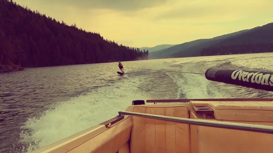 the image depicts a person waterskiing on a calm lake surrounded by mountains and lush green forests.  the view is from the back of a classicstyle motorboat, which is pulling the skier.  the water is relatively calm, with a noticeable wake trailing behind the skier. the sky is somewhat overcast, giving the image a slightly muted, vintage tone. a portion of a black tow rope, displaying the "overton's" brand, is visible in the foreground. the overall scene suggests a relaxing but active summer day spent enjoying water sports in a beautiful natural setting.