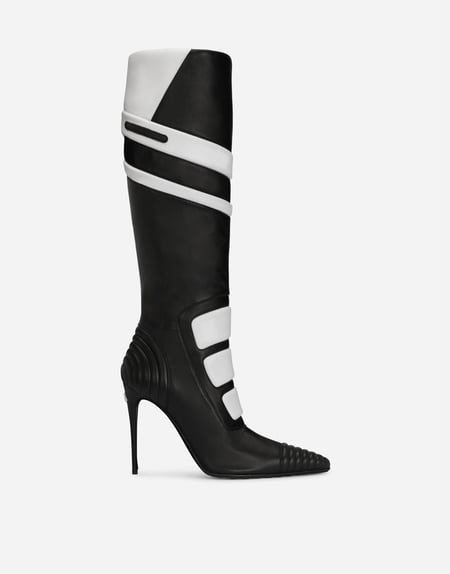 a black and white kneehigh heeled boot with a stiletto heel is shown against a white background. the boot is primarily black with white accents. the white accents are on the upper part of the boot, including a geometric design and several horizontal stripes. the heel is slender and tapered. the sole is textured near the toe. the background is plain and bright, placing emphasis on the boot.