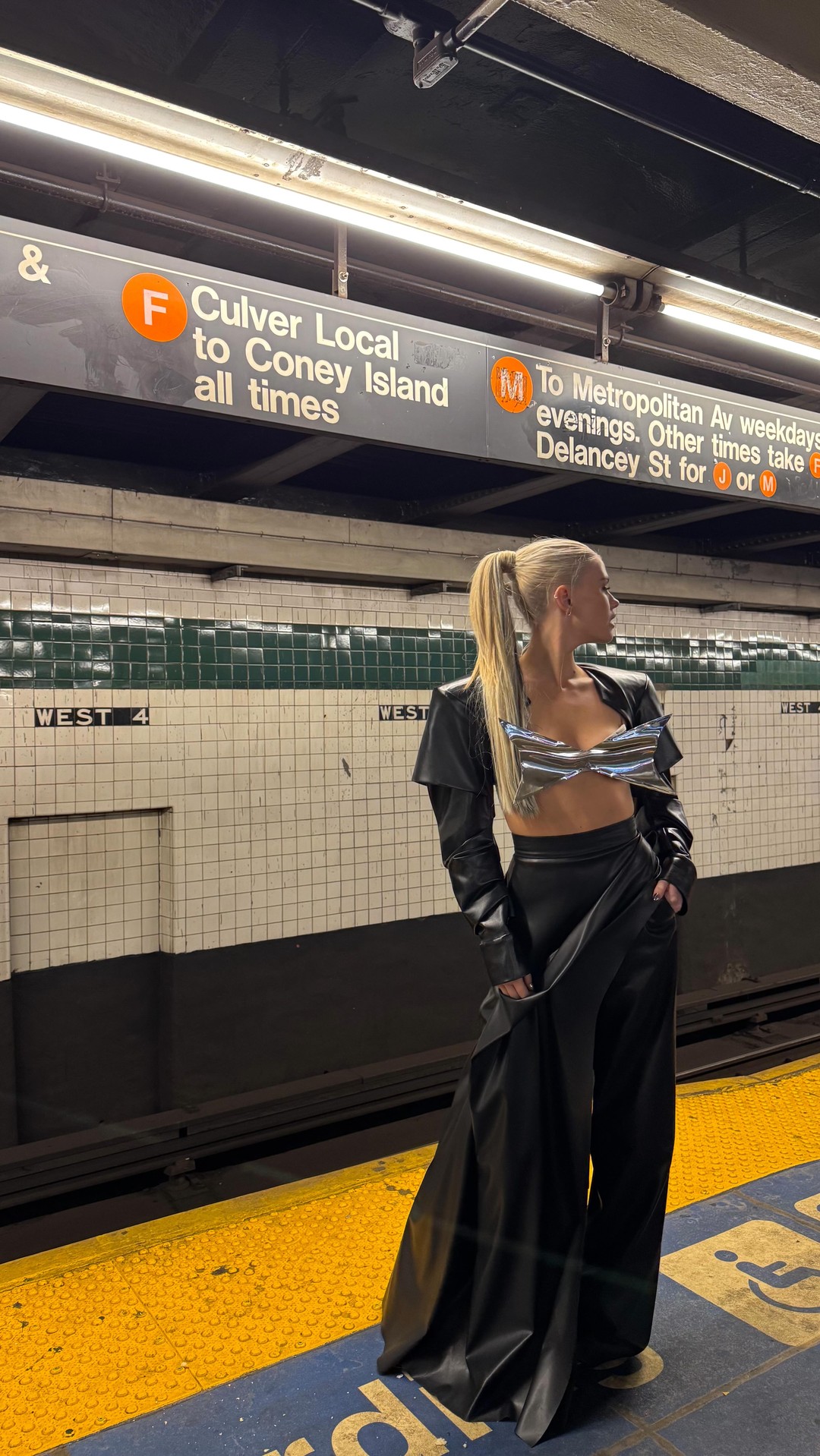 a woman with blonde hair in a high ponytail stands on a subway platform. she is wearing a black leatherlook outfit consisting of highwaisted wideleg pants, a cropped jacket, and a silver metallic bra top. above her head are signs for the f train (culver local to coney island) and the m train (to metropolitan ave evenings). the station tiles indicate "west 4" implying the west 4th street station.