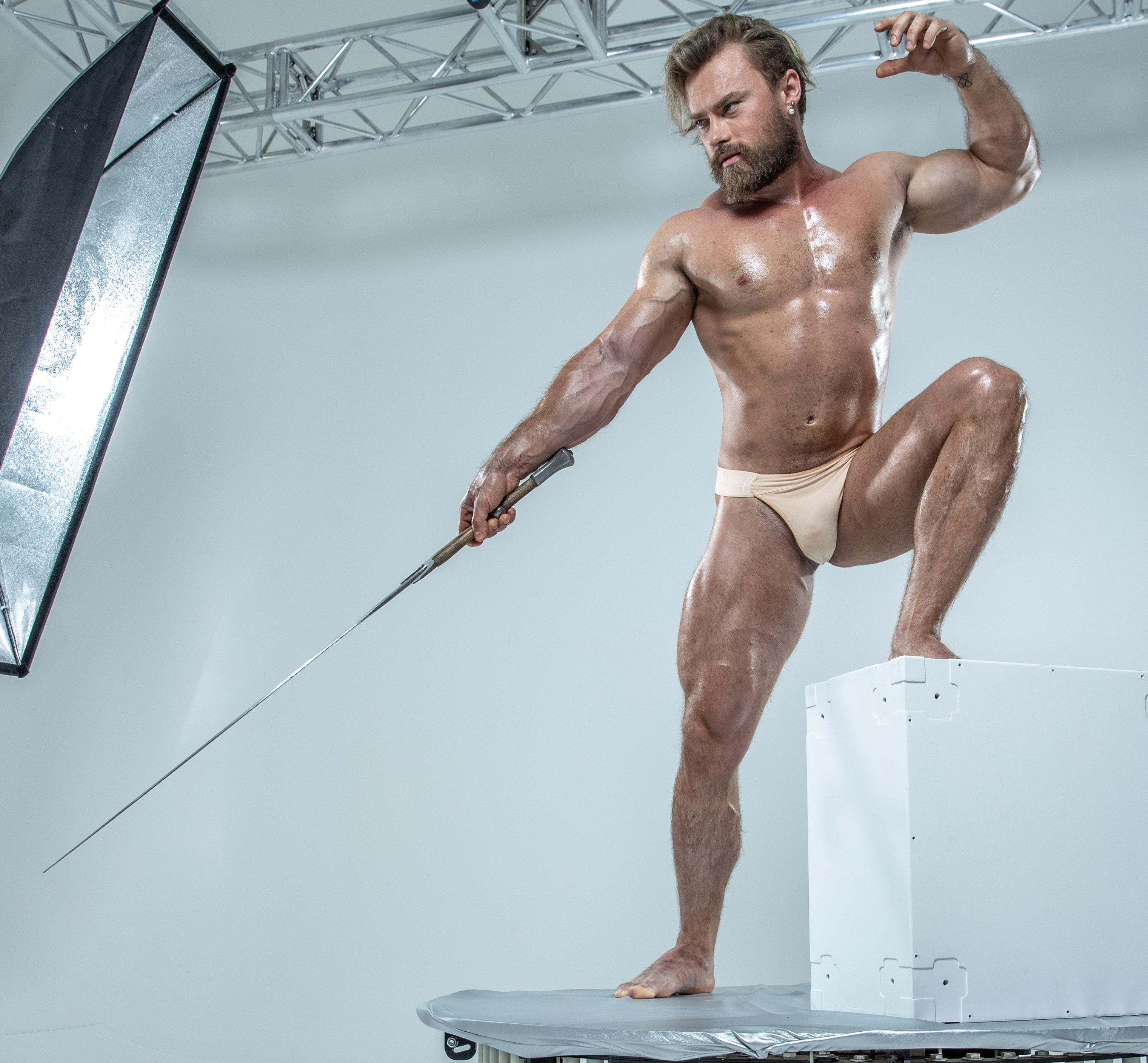 a muscular man with a beard is standing on a white platform in a photography studio. he is holding a fencing sword and is wearing only underwear. he is illuminated by artificial light, possibly from a softbox reflector on the left, creating a dramatic effect. the background is entirely white, focusing attention on the subject and his dynamic pose. the image exudes a sense of strength, power, and athleticism.