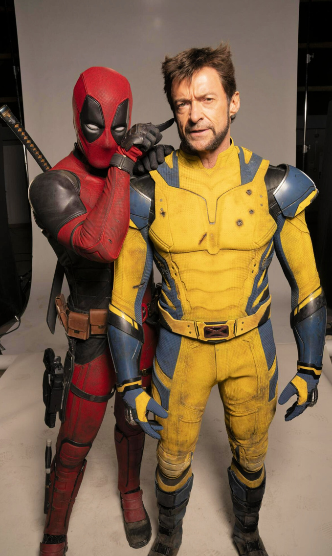 the image features deadpool and wolverine in costume, with hugh jackman portraying wolverine and ryan reynolds as deadpool. they are standing close to each other, with deadpool touching wolverine's ear. both characters are wearing their classic superhero costumes.