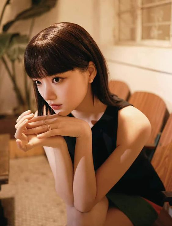 a young woman with dark hair and bangs is sitting on a chair in a cafe. she is wearing a black sleeveless top and a colorful skirt. she is resting her chin on her hands and looking off to the left side of the frame. the lighting is warm and natural, creating a soft and inviting atmosphere.  the image has a sense of intimacy and evokes a feeling of contemplation.