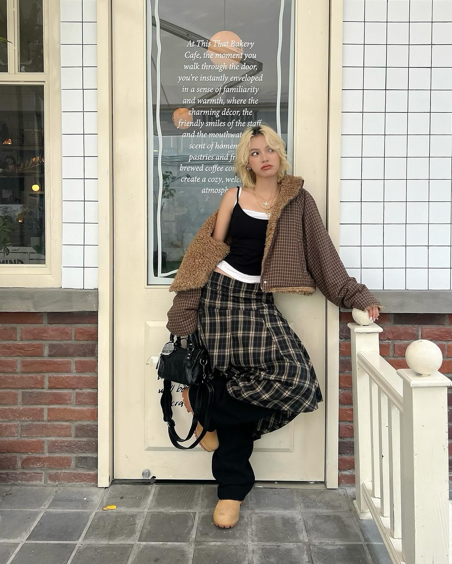 a young woman, possibly a model, is posing outside a bakery café. she's wearing a stylish outfit that includes a brown teddystyle jacket over a black knit top and a brown and beige plaid pleated skirt.  she also has on black, wideleg trousers underneath the skirt. brown boots and a black bag complete the look. she's leaning against a white railing and her pose is relaxed but fashionforward.  the bakery's window display is visible, showcasing a text message that describes the café's welcoming atmosphere.  the overall aesthetic is a blend of vintage and contemporary styles, with a hint of korean fashion influences.  the setting appears to be in a stylish urban area.