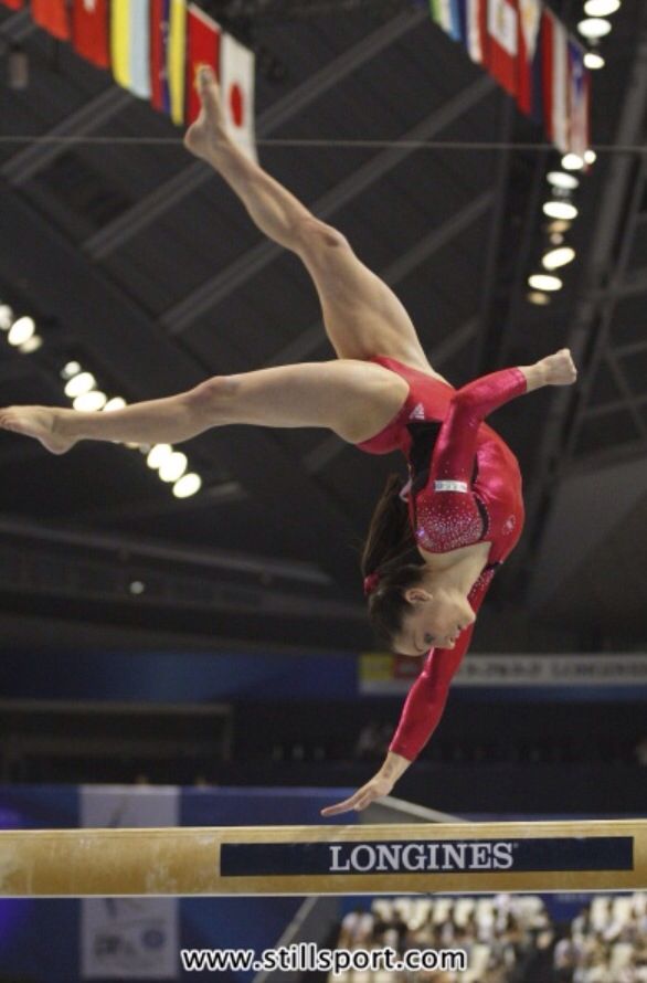a female gymnast performs a back handspring on the balance beam, midair with a red leotard, during a gymnastics competition. the background shows a blurred view of an audience, spectators, and flags.