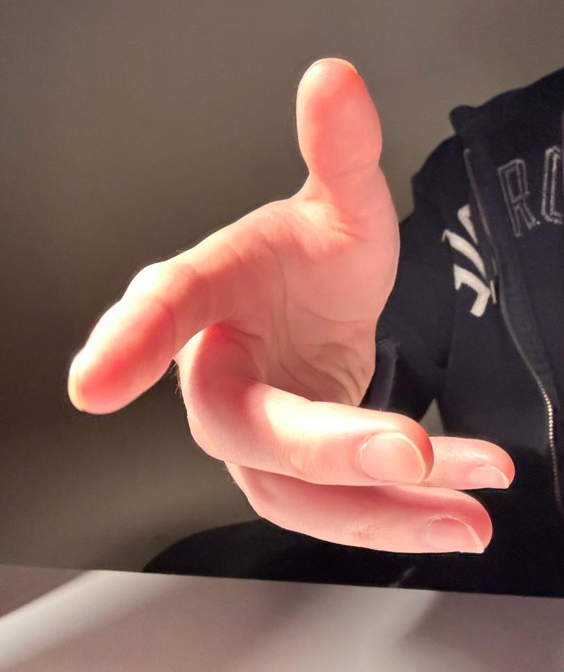 the image shows a closeup view of a person's right hand, partially illuminated by artificial light, reaching out towards the viewer. the hand is pale and has long fingers.  the background is dark and out of focus, with a portion of a black jacket visible. the hand is in sharp focus, with the light creating strong shadows.