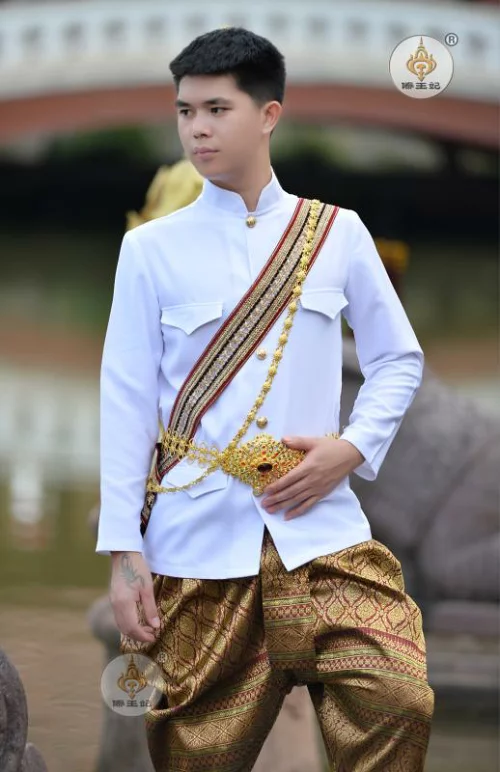 a man is wearing traditional thai clothing. he is wearing a white shirt, gold sash, and patterned pants. he is standing outside in front of a lush green background.