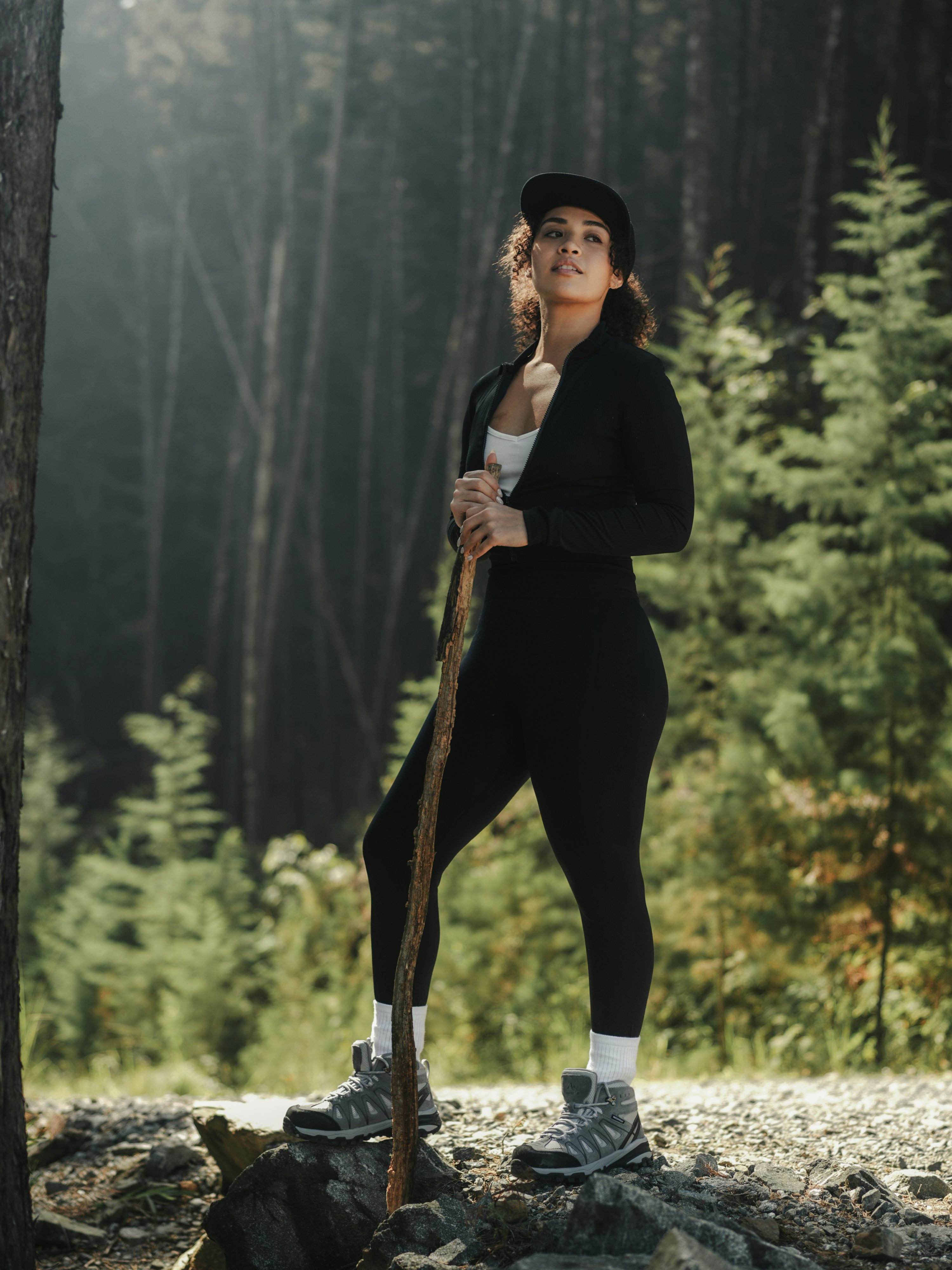 a woman in a black athletic outfit stands in a forest holding a walking stick. she is wearing leggings, a jacket, a baseball cap, and hiking boots. she is standing on a rocky outcrop. the forest is dense with trees in the background.