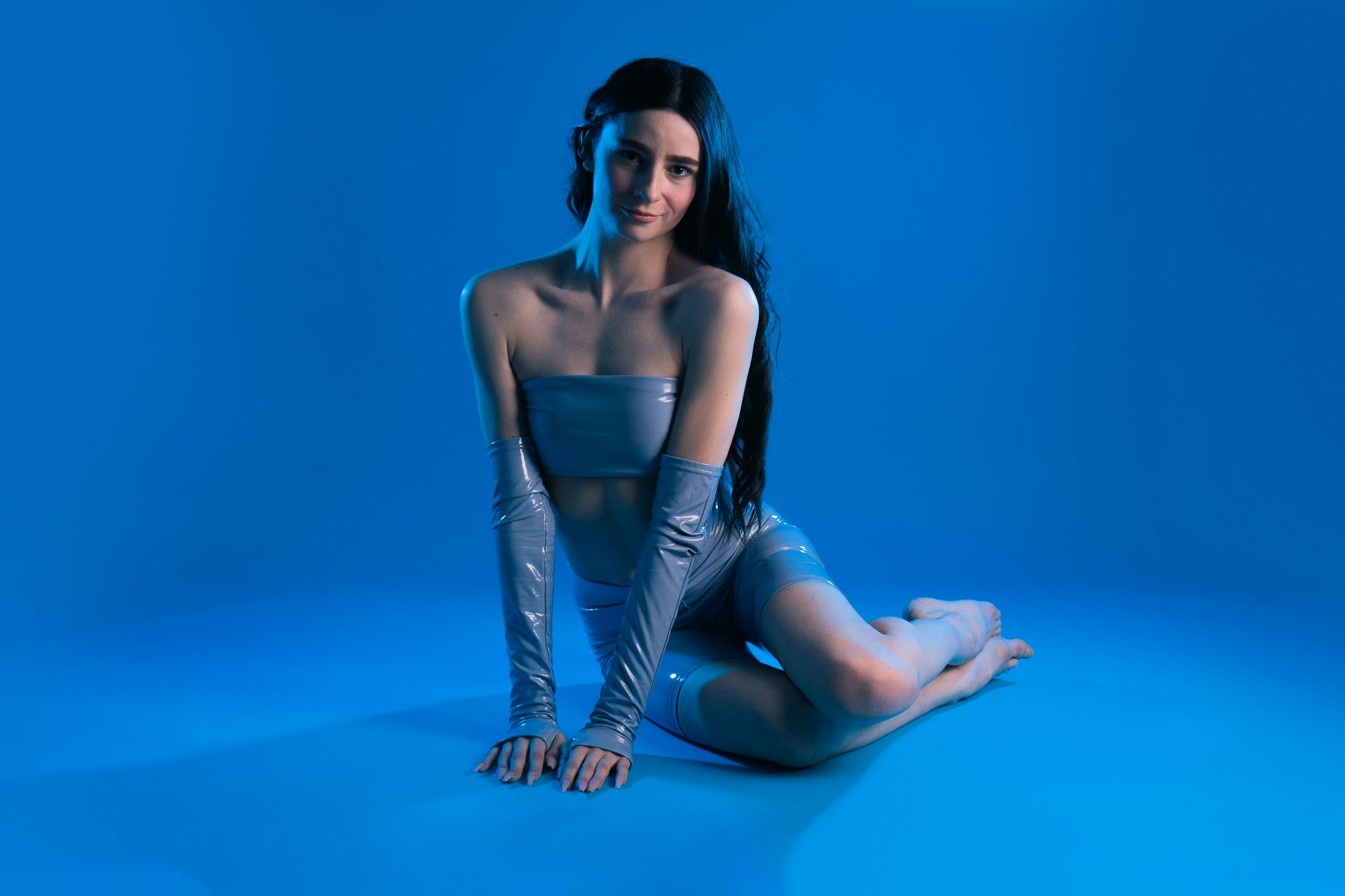 a young woman with long dark hair is sitting on a blue background. she is wearing a shiny, light blue latex bodysuit that includes a strapless top and short shorts. she also has matching long gloves. the image is stylized with blue lighting, creating a futuristic or cyberpunk vibe. the model is posing confidently, with a direct gaze towards the camera.  the overall aesthetic is sleek, minimal, and fashionable.
