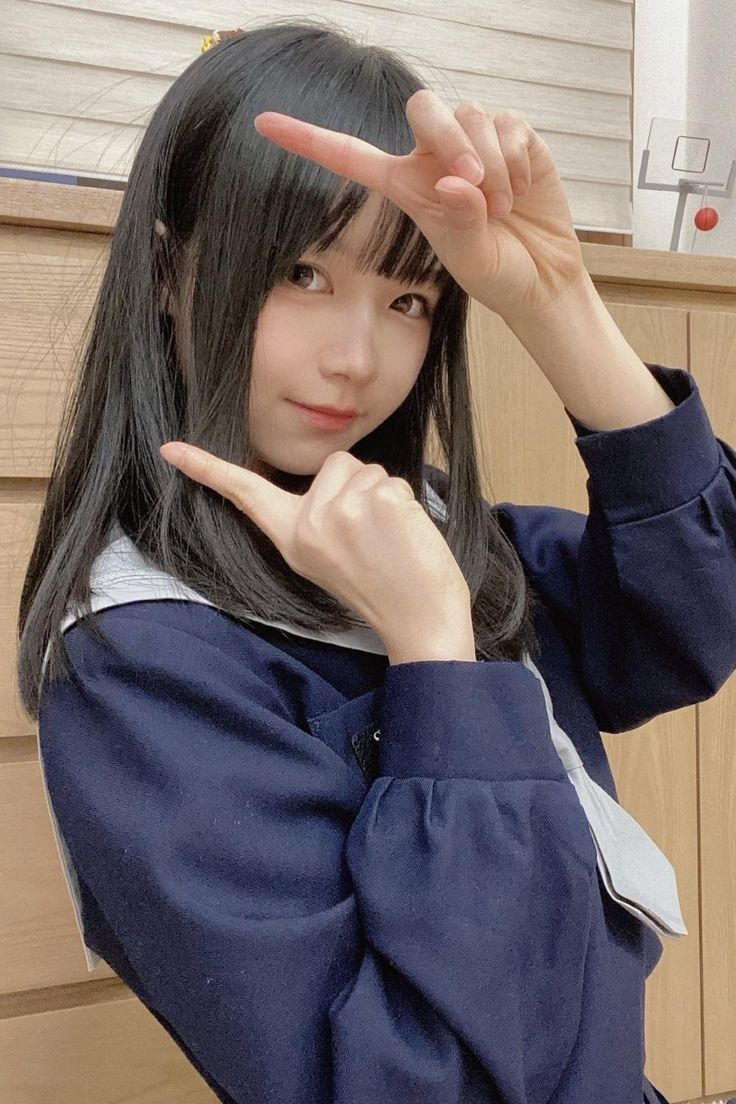 a young woman with dark hair and bangs is posing for a selfie. she is wearing a japanesestyle school uniform with a sailor collar. she is indoors and lit by artificial light. she is making a framing gesture with her hands.