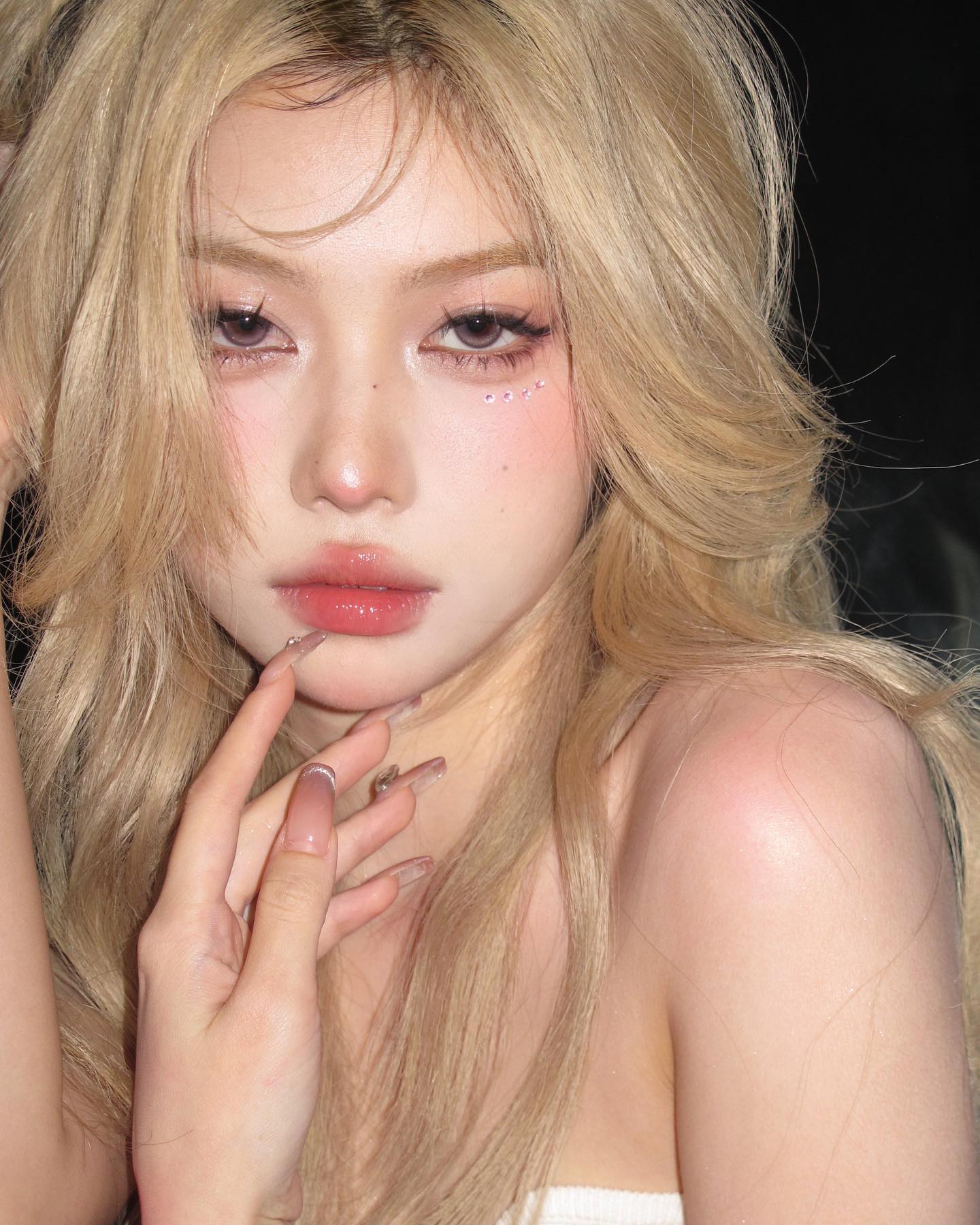 a closeup portrait of a young asian woman with blonde hair. she has immaculate makeup with a focus on her eyes and lips. the image highlights current beauty trends.  she is touching her face with her hand, which has long, decorated nails. the lighting is soft, giving her skin a warm glow.