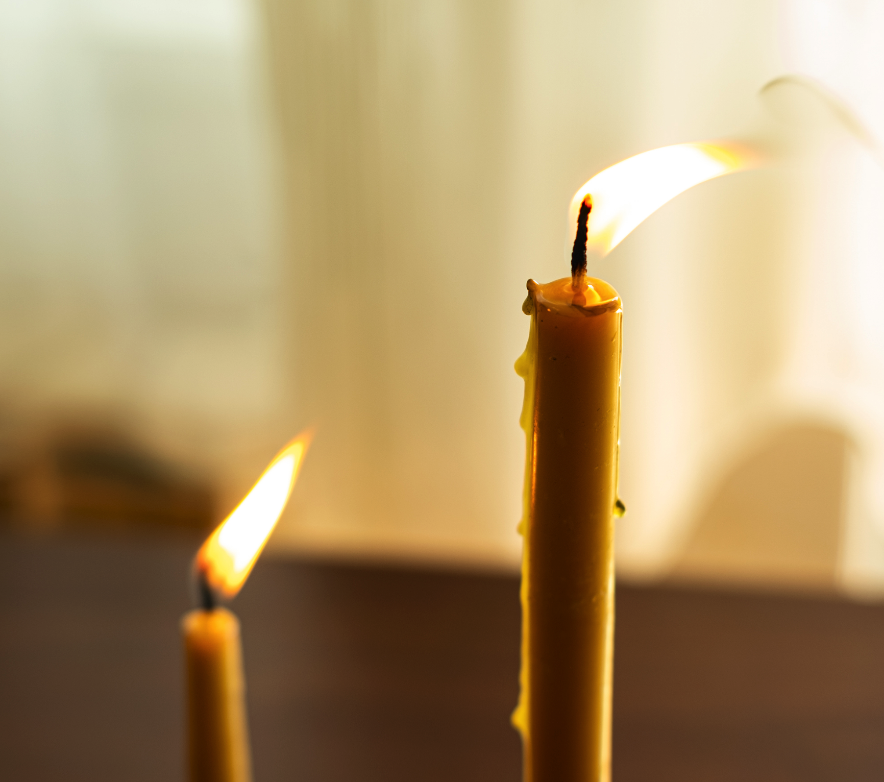 a closeup shot of two candles, one with a lit flame and the other with a melted wax pool. the candles are yellow and stand upright against a blurred background. the image evokes a sense of peace and tranquility, perhaps reflecting on spirituality or remembrance.