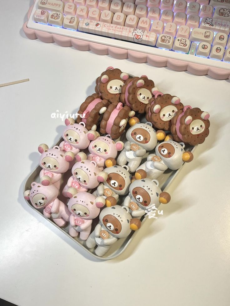 this image showcases a keyboard with pink accents and decorative keycaps, positioned above a silver tray filled with various rilakkumathemed items. the tray contains small rilakkuma figures, some wearing pink hoods and others wearing white hoods, as well as rilakkumashaped sandwich cookies with pink filling. the overall aesthetic is cute and kawaii.