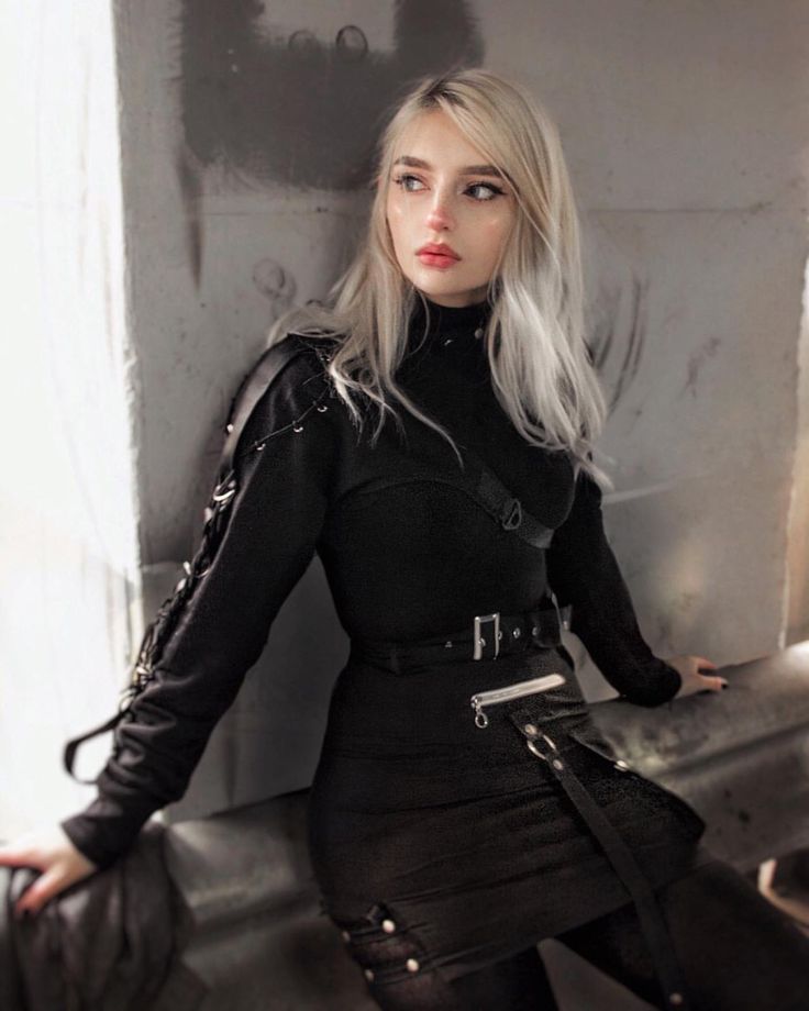 the image shows a young blonde woman with pale skin, wearing a complete black outfit in a gothic style.  the outfit consists of a longsleeved turtleneck top with detailed straps and buckles on the shoulders and sleeves,  a black miniskirt with additional straps and buckles around the waist and hemline, and a matching black belt.  the woman is leaning against a grey, textured wall, looking off to her side with a serious, almost melancholic expression. the overall style is dark, edgy, and fashionforward, with a hint of cyberpunk influence. the lighting is moody and emphasizes the textures of both the clothing and the wall.