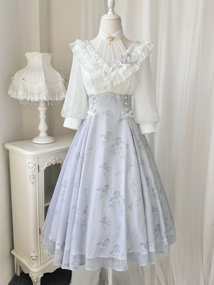 a fulllength view of a light purple lolita fashion dress displayed on a mannequin. the dress features a white lace blouse with a high neckline and ruffled trim, paired with a highwaisted skirt adorned with a delicate floral and bird print. the skirt has a layered, flowing silhouette, adding to the dress's elegance. the background showcases a glimpse of a room with a white wall and a vintagestyle nightstand.
