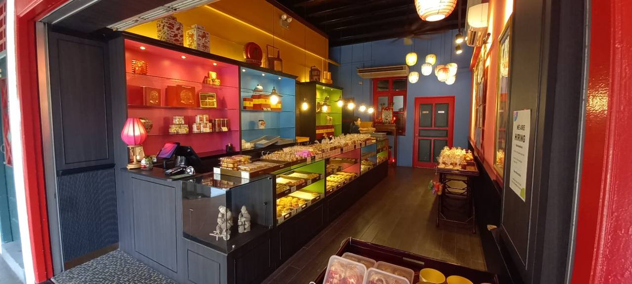 a small bakery shop with colorful shelves filled with sweets and pastries. the shop has a red, yellow, blue, pink and green color scheme and is decorated with chinese lanterns. there is a cash register and counter to the left of the image. there is a sign on the right side of the image that says "we are hiring". the shop has a glass storefront and window on the left side. the doorway to the shop is on the left side of the image. there are decorative details and trim on the shop’s interior.  the image is taken from the inside of the shop, looking out toward the doorway.