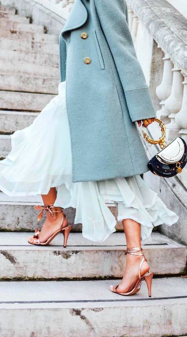 the image shows a stylish woman ascending a stone staircase. she's wearing a light blue, doublebreasted peacoat over a flowing, lightcolored pleated midiskirt.  her footwear consists of rose gold anklestrap heels. she carries a chloé handbag with a distinctive circular gold top handle. the overall style is elegant and chic.  the setting appears to be outdoors, with the emphasis on the woman's fashion.