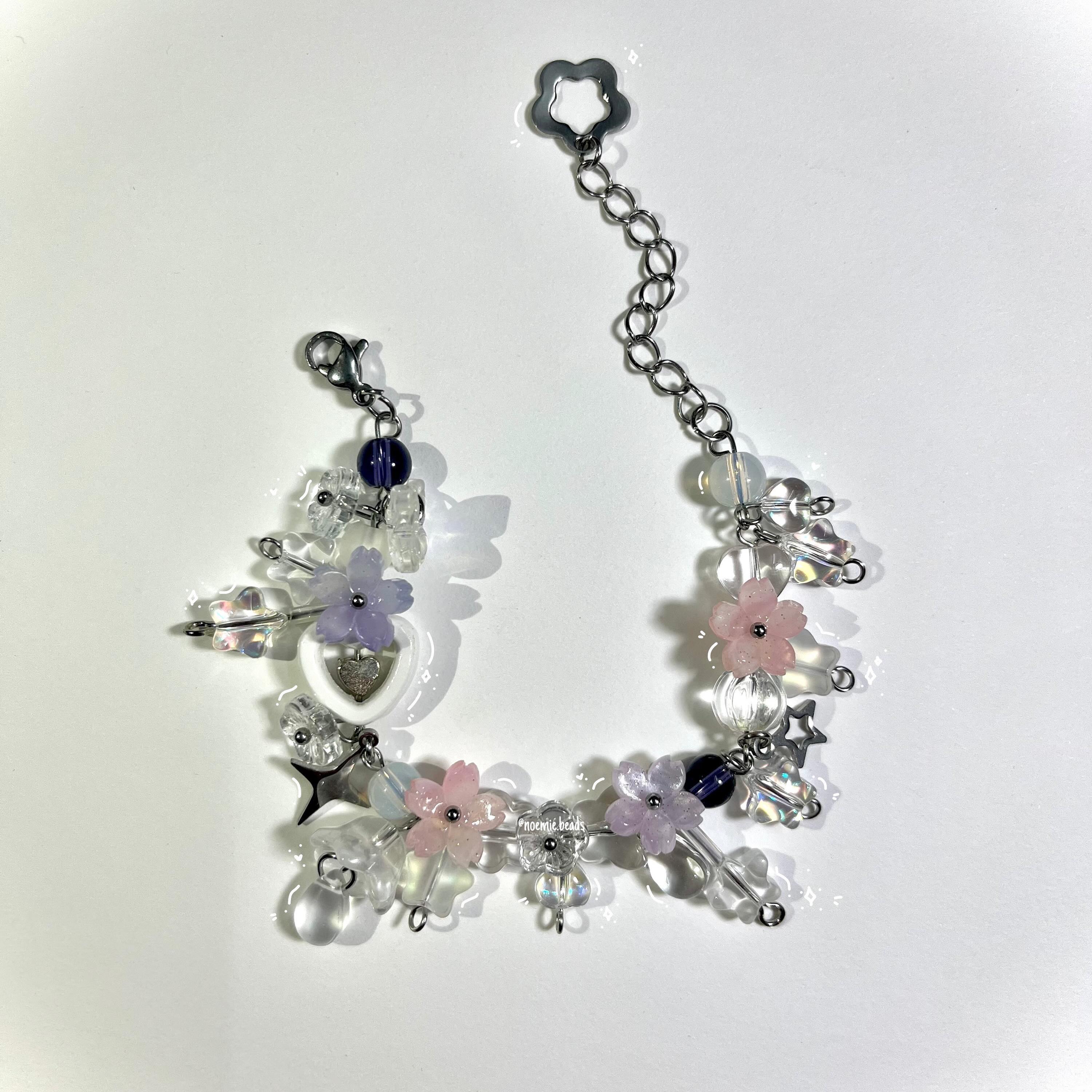 a handmade bracelet lies on a white surface. the bracelet is made with an assortment of clear, pink, and purple flower beads, with metal star and heart charms, and various clear and dark blue beads. it is connected by a metal chain and clasp.