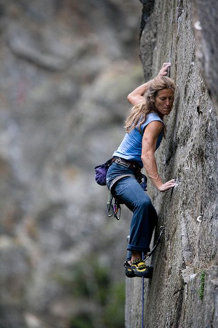 a woman rock climber scales a large rock face. she is wearing a harness and climbing gear. she is focusing on the next hold and is determined to make her way to the top. the rock face is smooth and there is no easy path for her to take. this is a challenging climb, but she is up for the challenge.