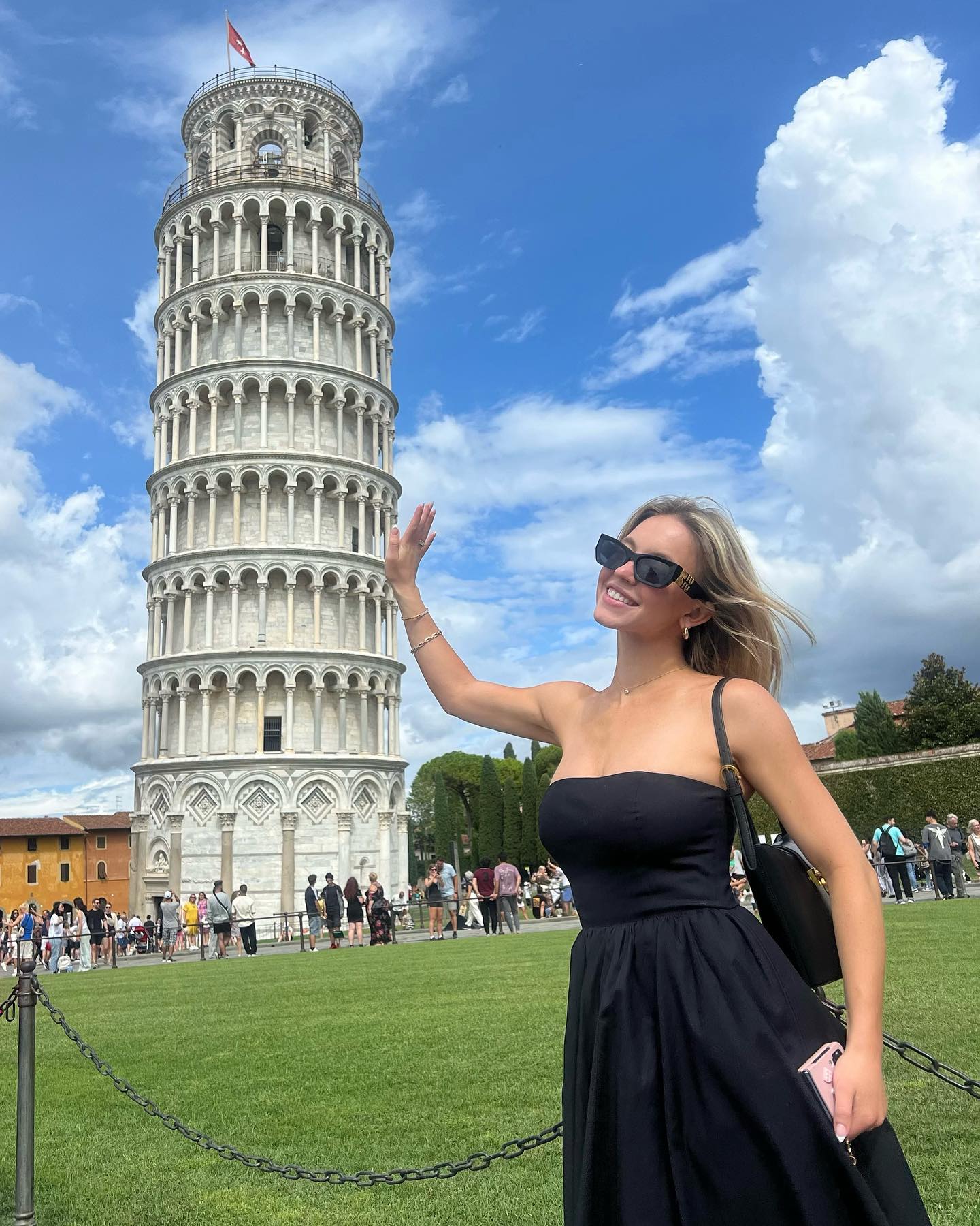 a woman in a black strapless dress is standing in front of the leaning tower of pisa, waving her hand. she is wearing sunglasses and smiling happily. the sky is bright blue with fluffy white clouds. there are other tourists visible in the background. the scene is picturesque and captures the essence of a joyful summer vacation in italy.