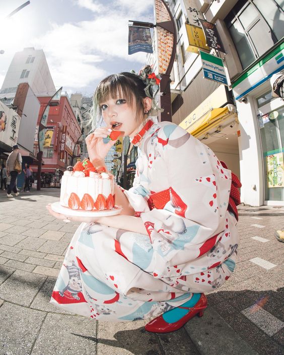 a young woman wearing a playful modern yukata is squatting on a busy city street, holding a whole strawberry shortcake. she's about to take a bite of the cake, looking directly at the camera with a mischievous smile. the wideangle lens captures the dynamic energy of the urban environment.