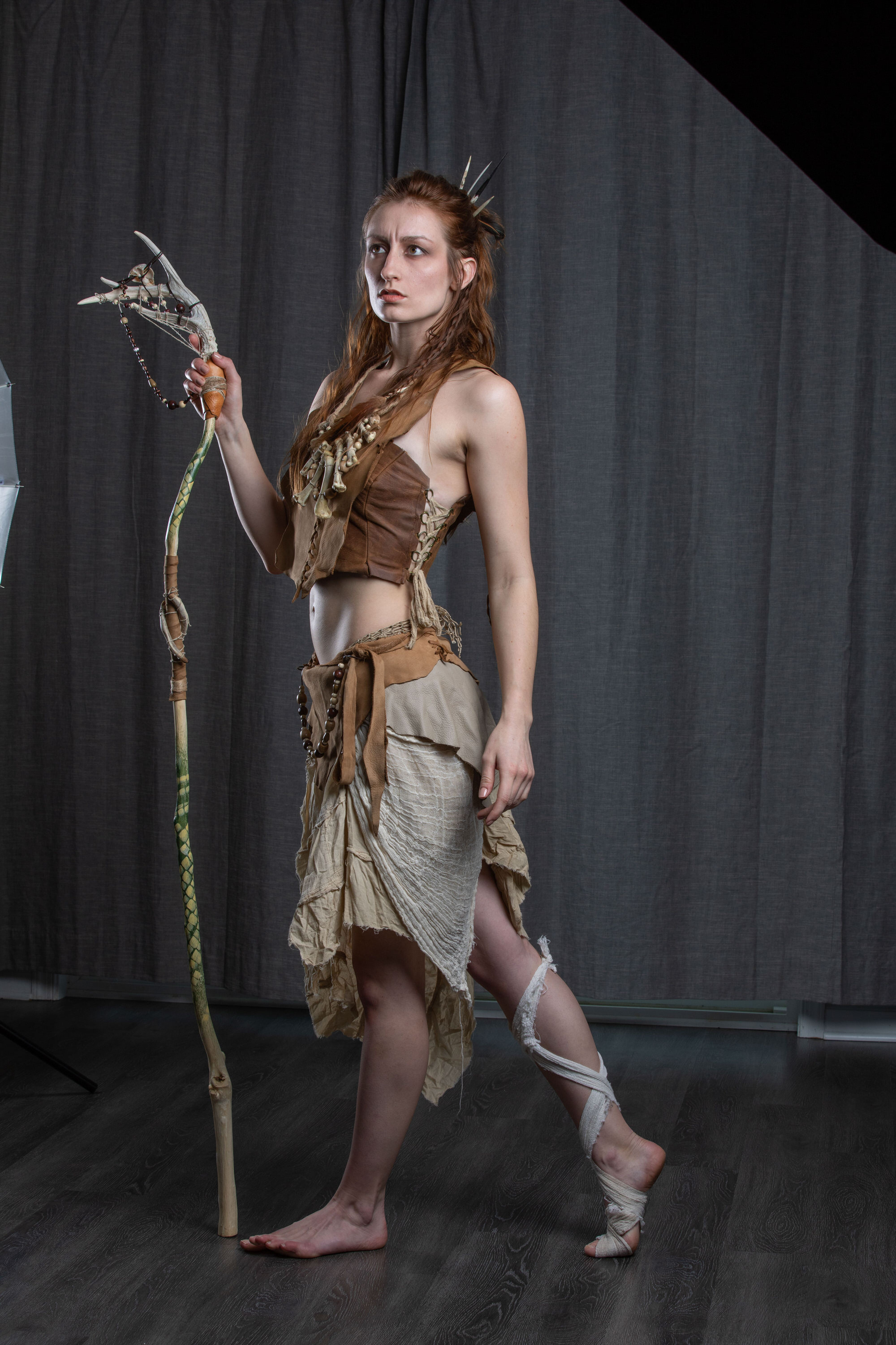 a young woman with reddish hair styled in an elaborate braid and adorned with bones stands confidently against a dark gray backdrop. she is dressed in a tribalinspired costume, featuring a leather top, a flowing beige skirt, and bone and leather accessories. her left leg is partially wrapped in fabric, and she holds a long, gnarled staff topped with antlers. her attire and the staff suggest a fantasy character, perhaps a warrior or a mystical figure.  the studio setting is evident from the partially visible lighting equipment in the background.