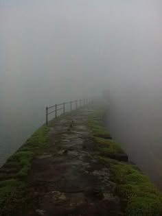 a path on a stone bridge stretches into the distance, shrouded in a thick fog. the railing on the side of the path is visible, but everything else is obscured by the dense mist. the path is lined with grass and moss, and the overall feeling of the image is one of tranquility and mystery.