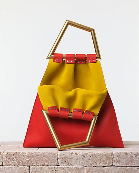 a close up of a bright red and yellow leather handbag with a gold geometric handle. the bag is sitting on a brick surface.