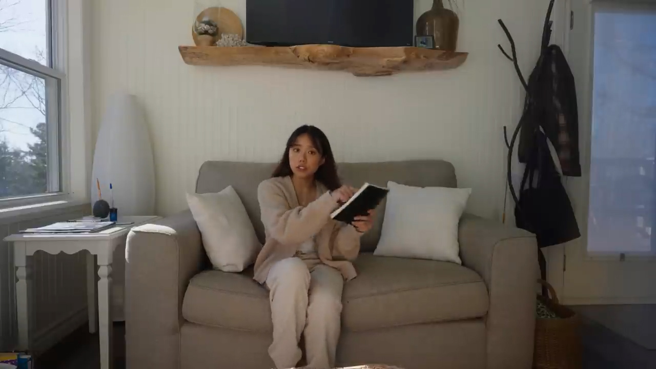a young woman sits on a beige couch in a cozy living room, bathed in soft, natural light streaming in from a window to her left.  she is engrossed in a book held open in her hands. the room is minimally decorated in neutral tones, with a wooden shelf above the couch and a white side table to the left. a woven basket sits on the floor beside the couch. the overall atmosphere is one of calm and relaxation.