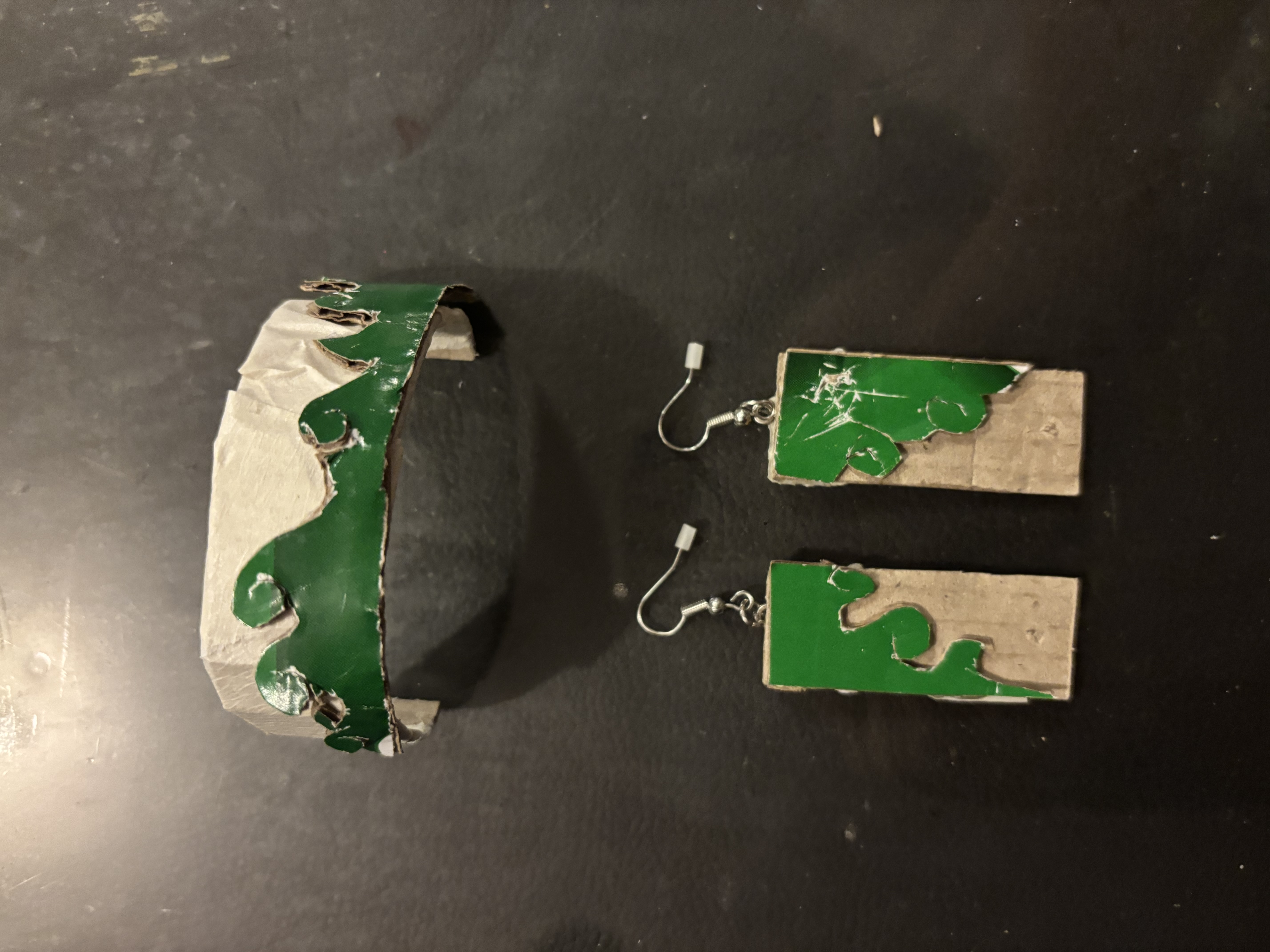 the image shows a handmade set of jewelry. there are a pair of earrings and a crown. the earrings are rectangular cardboard pieces with an irregular green design on the top half and silver ear hooks attached. the crown is made of cardboard wrapped in white tape, with a green wavy design along the front edge.