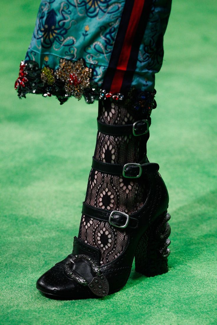 closeup shot focuses on a model's leg and foot, showcasing a pair of unique black highheeled shoes. the shoes are adorned with multiple buckles and have a textured, possibly leather, upper. the model is wearing black lace stockings that are visible above the shoes. the lower part of the model's outfit is also visible, which features an intricately designed teal and red patterned fabric with decorative embellishments along the hemline. the background is a vibrant green surface. the overall aesthetic suggests a fashion show or runway setting. the image highlights the detail and texture of both the shoes and the clothing.