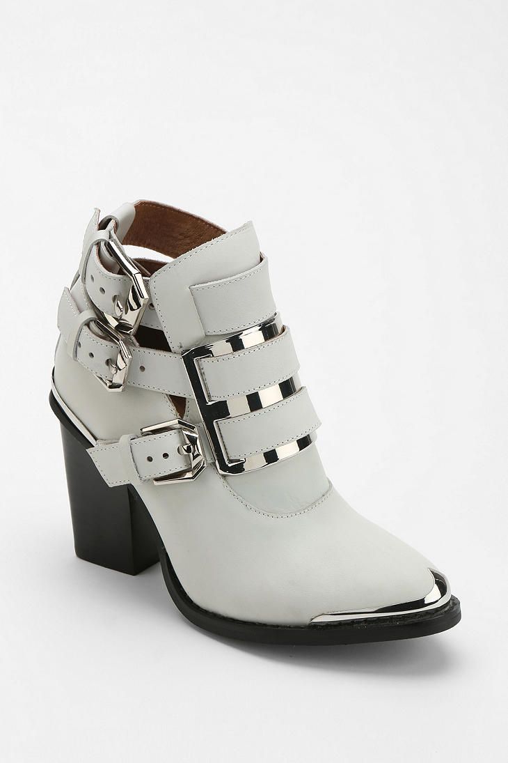 a single light grey leather heeled boot is displayed against a white backdrop. the boot has a black block heel, a pointed toe with silver metal caps, and three straps across the front of the ankle, each with a silver metal buckle. the straps have silver metal accents.