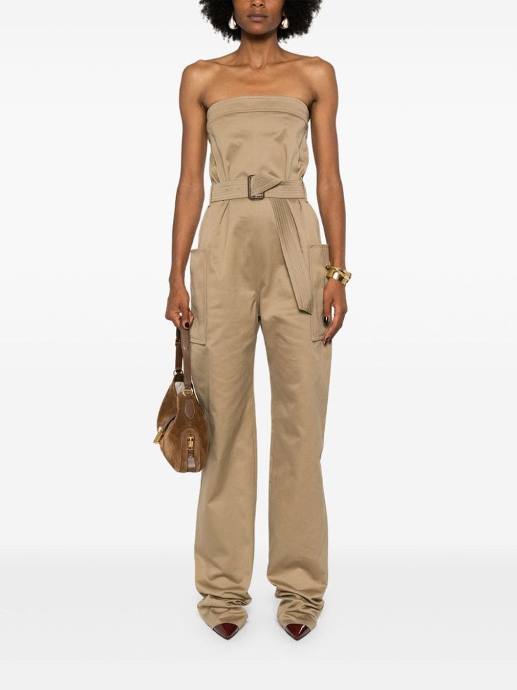 a woman wearing a strapless beige jumpsuit with a brown belt and a brown handbag. she is posing in a studio with a white background.
