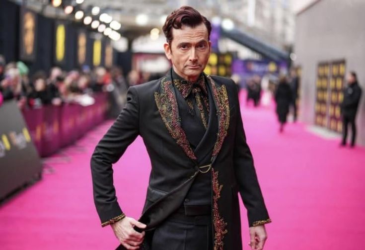 a fulllength shot of david tennant standing on a pink red carpet. he's wearing a black suit with intricate embroidery along the lapels, cuffs, and collar. he's holding open the side of his suit with his left hand. in the background, there are other people and event lighting.