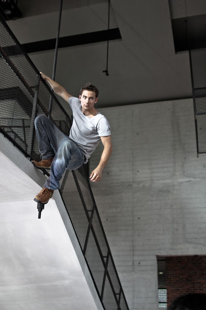 a man wearing a gray tshirt and jeans is climbing on a black metal staircase indoors. he is wearing brown shoes and appears to be doing parkour or an urban adventure activity. the background features a concrete wall and the setting looks like an industrial or modern building.