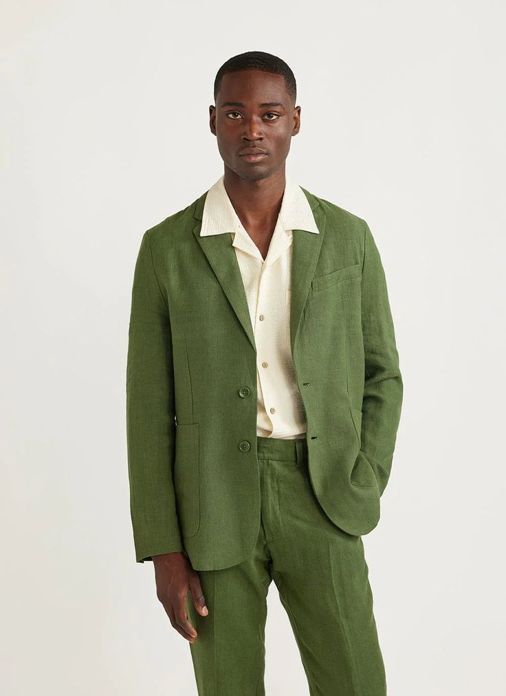 a young man in a green linen suit, with a white linen shirt underneath, poses for the camera. he has dark hair and a serious expression.