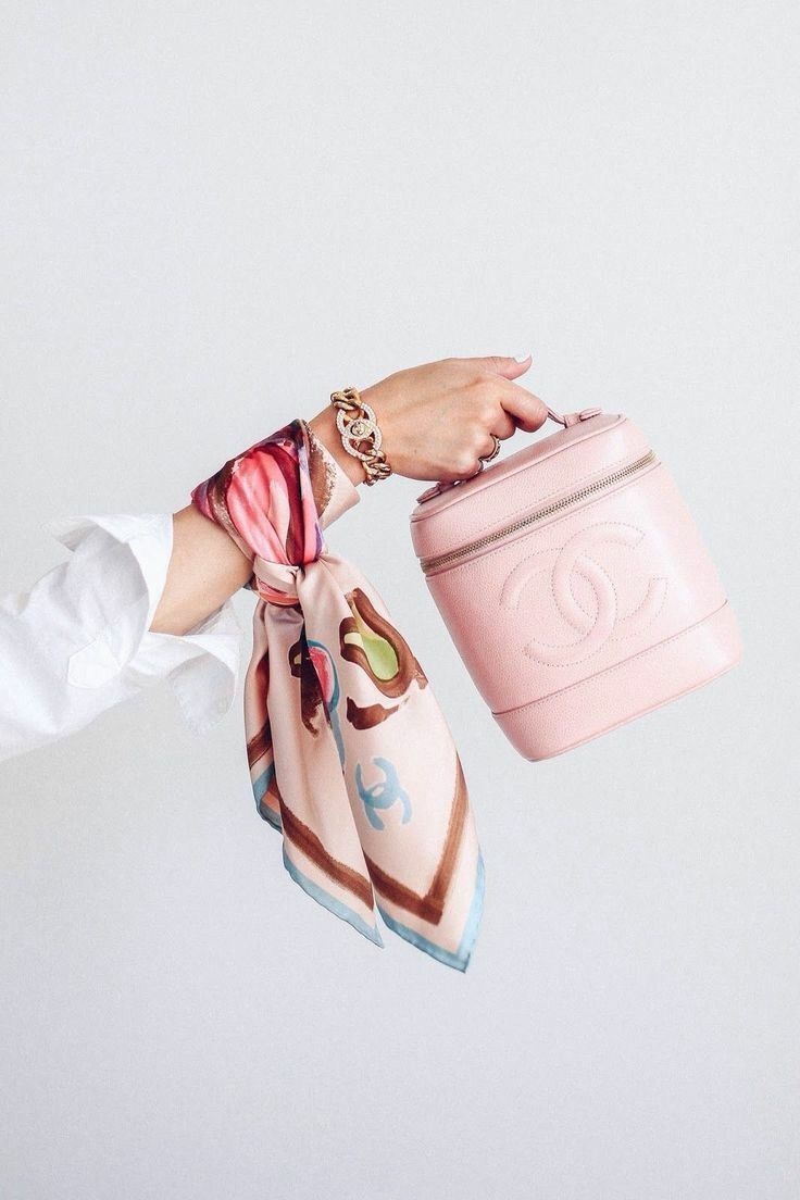 a closeup image of a woman's arm adorned with a pink silk scarf and a gold chanel bracelet. she is holding a pale pink chanel vanity case.  the background is a simple, light gray.  the overall feel is elegant and sophisticated, showcasing highend fashion accessories.