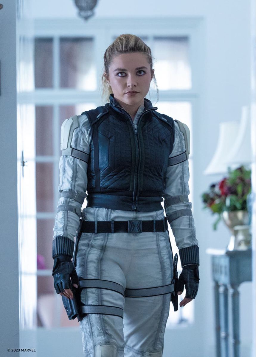 a woman dressed in a black widow costume is standing in a doorway, looking intensely at something offscreen. the room behind her is white and has a window to the right, along with a table with a vase of flowers. the woman is wearing a black vest over a white jumpsuit, with black gloves and a gun on her hip.