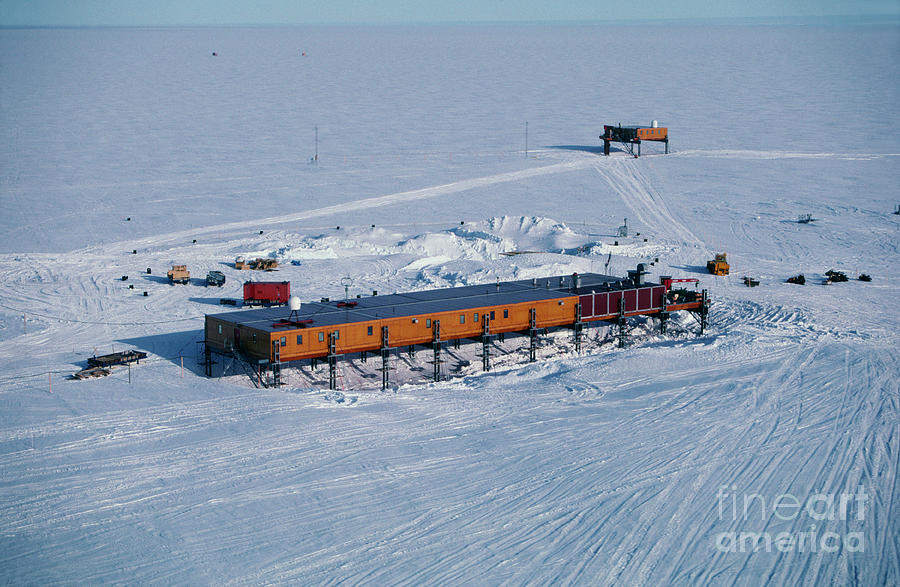 an aerial view of a research station in antarctica, the station is built on stilts with a long, narrow, building, and a few smaller structures.  the surrounding area is covered in snow and ice, with tire tracks from vehicles. the sky is blue and clear, and the horizon is distant.
